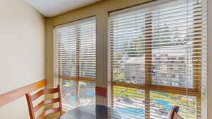 Un balcón con mesa y sillas, ventanas amplias con persianas y vistas a una piscina y edificios.