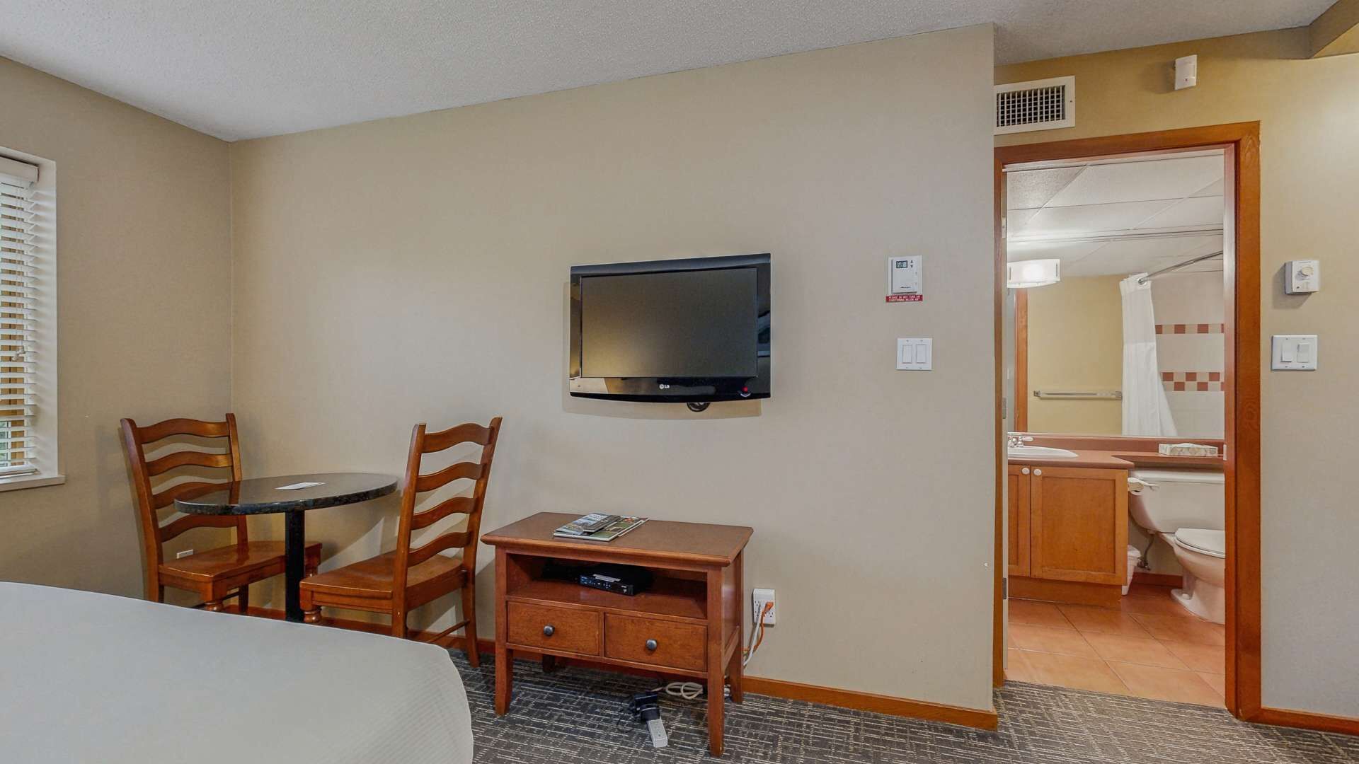 A hotel room with a bed, a TV mounted on the wall, a desk with a chair, and a bathroom visible through an open door.