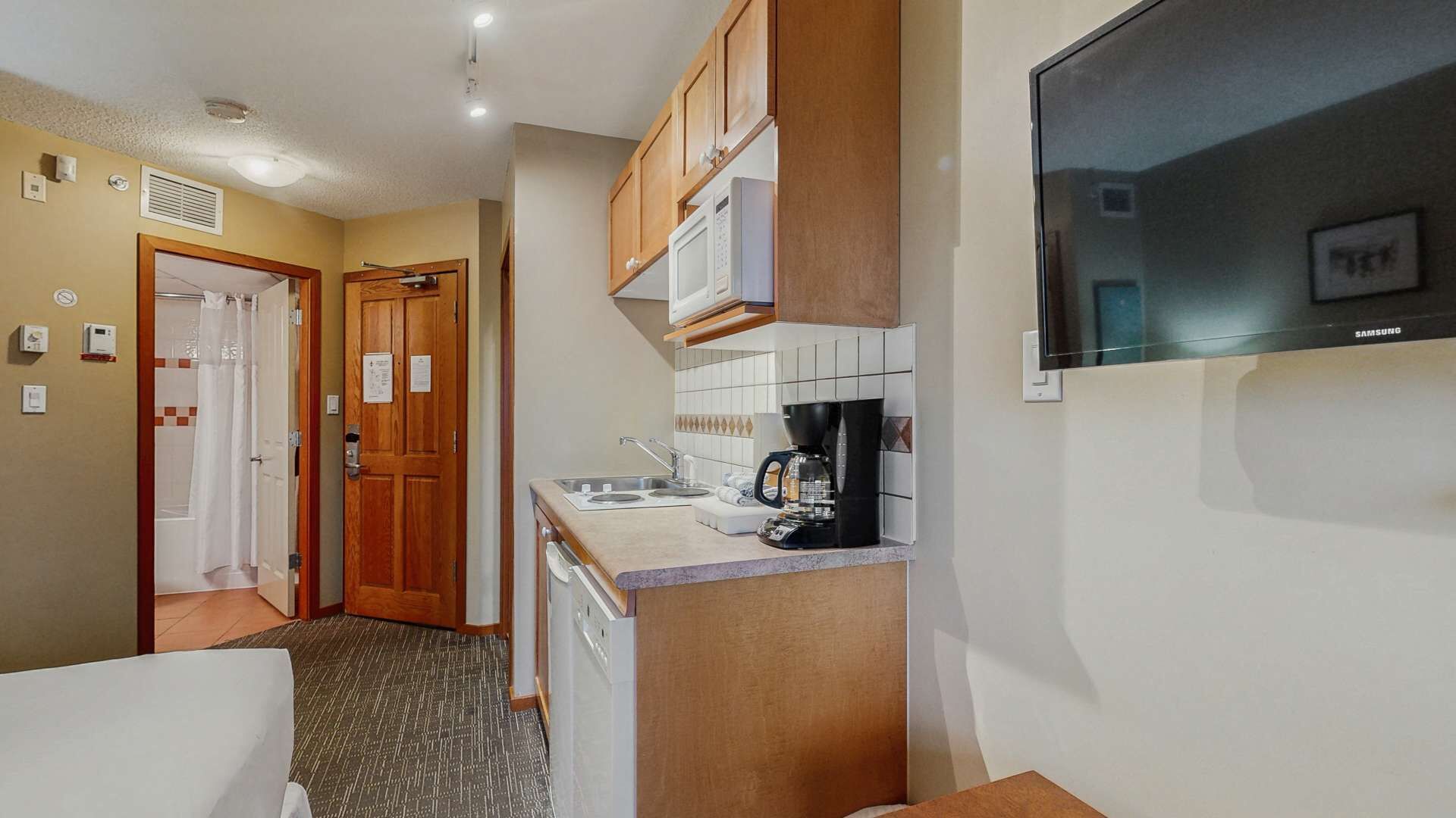 Una cocina compacta con armarios de madera, un microondas, una cafetera y un televisor Samsung montado en la pared.