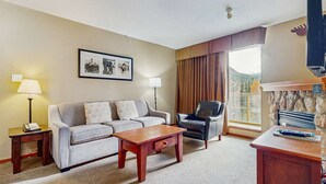 Une chambre d’hôtel avec un canapé, un fauteuil, une petite table, une lampe, un tableau encadré, une fenêtre avec des rideaux et une cheminée en pierre.