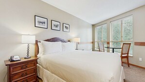 Une chambre d’hôtel avec un grand lit, deux tables de chevet avec des lampes, une petite table en bois, une chaise et trois tableaux encadrés accrochés au mur.