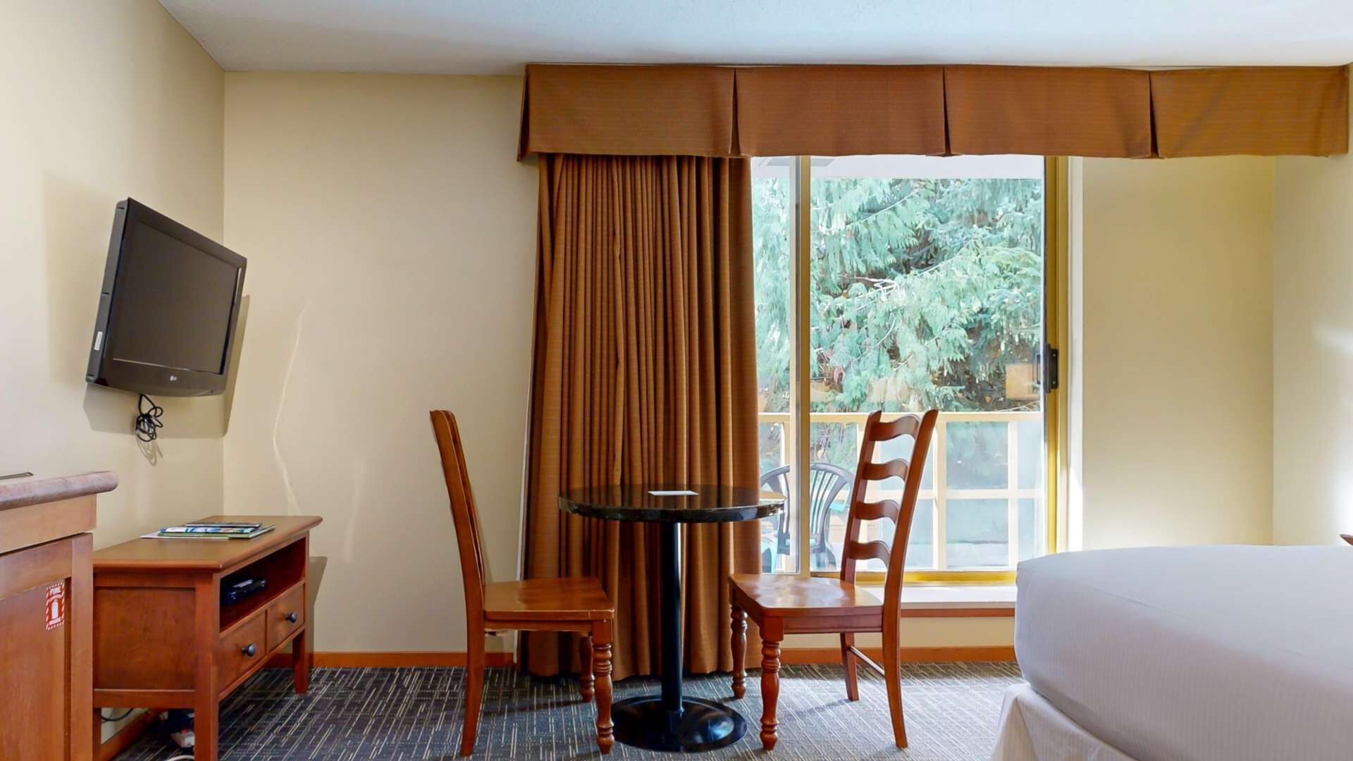 Une chambre d’hôtel comprenant un lit, un bureau avec une chaise, une petite table avec une chaise, un téléviseur fixé au mur et une fenêtre avec des rideaux.