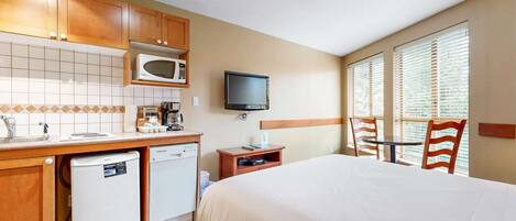 A hotel room with a bed, a small kitchenette, a television, and a window with blinds.