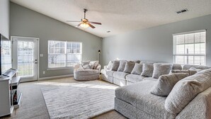 A spacious living room with a large sectional sofa, a round ottoman, and a ceiling fan. There are two windows with blinds, a door, and a small table with a lamp. - Work-friendly Madison Home Near Research Park (Madison)