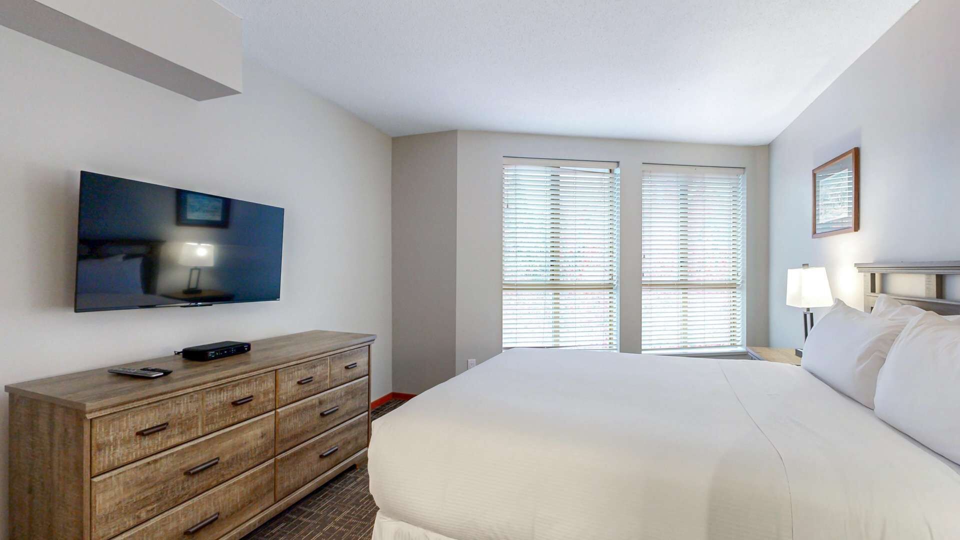 Een hotelkamer met een bed, een aan de muur bevestigde televisie, een houten dressoir en een raam met zonwering.