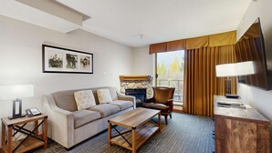 Une chambre d’hôtel avec un canapé, une table basse en bois, un fauteuil, une cheminée, un téléviseur et un tableau encadré au mur.