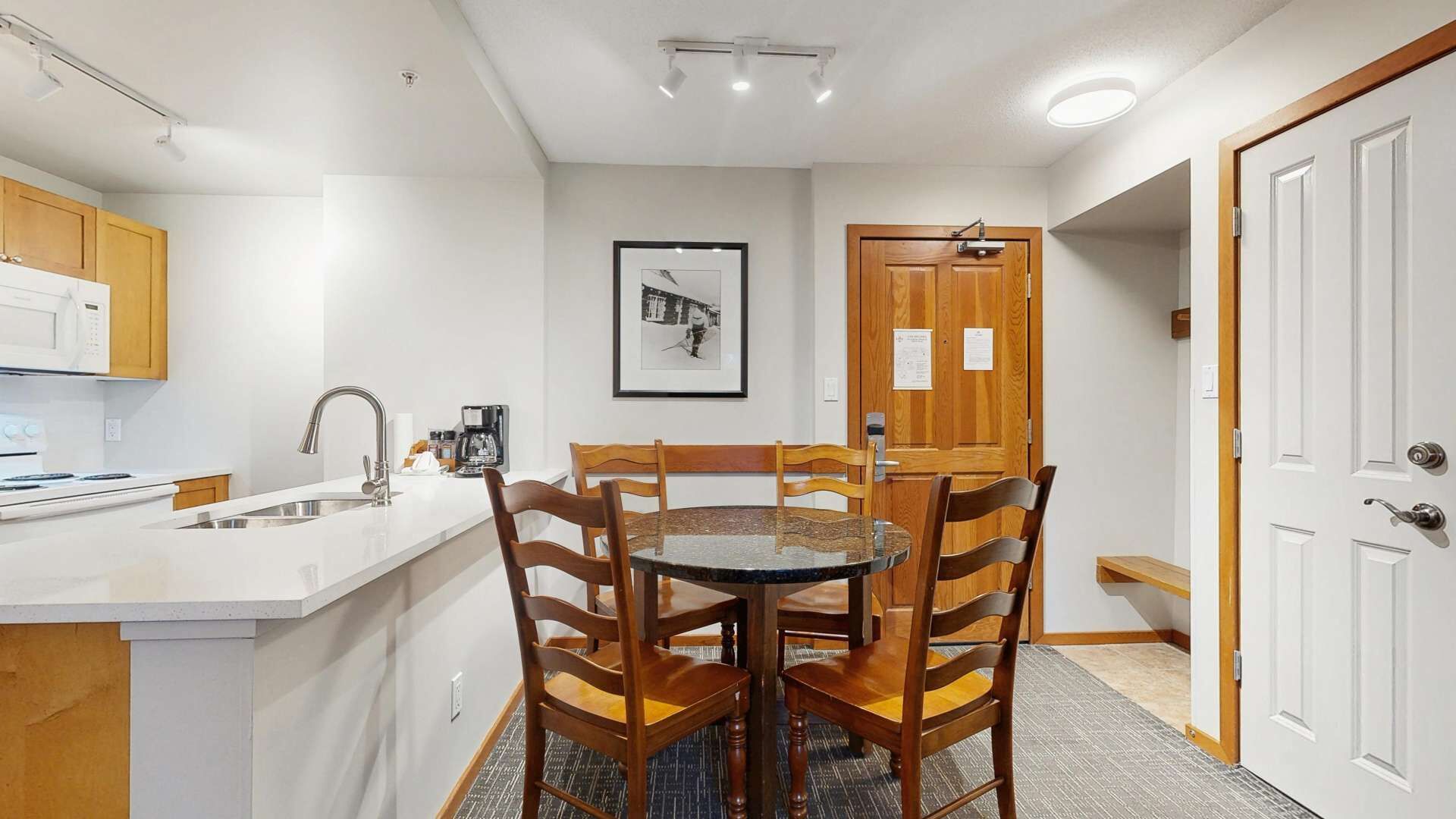 Een kleine keuken met een wit aanrecht, een spoelbak en een koffiezetapparaat. Er staat een eettafel met vier houten stoelen en aan de muur hangt een lijst met een afbeelding.