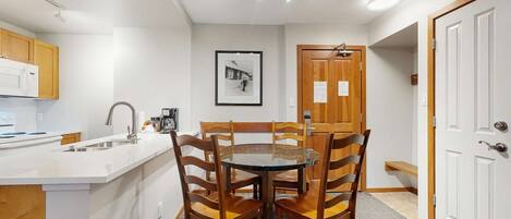 Een kleine keuken met een wit aanrecht, een spoelbak en een koffiezetapparaat. Er staat een eettafel met vier houten stoelen en aan de muur hangt een lijst met een afbeelding.