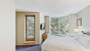 Ein Hotelzimmer mit einem großen Bett, einem Fernseher, einem Schreibtisch und einem Fenster mit Jalousien.