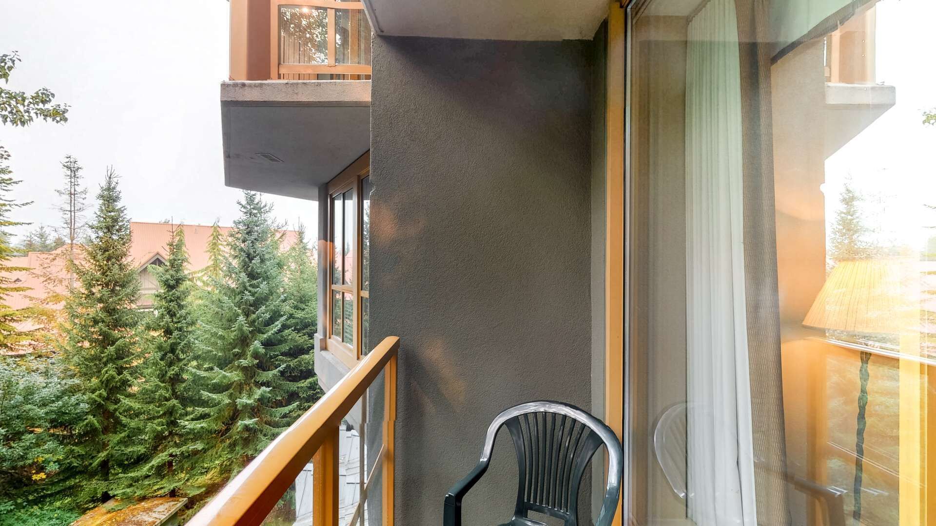 A balcony with a view of evergreen trees, a chair, and a building with a balcony.