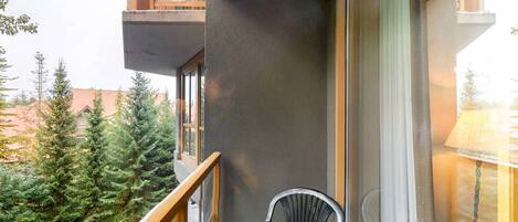 A balcony with a view of evergreen trees, a chair, and a building with a balcony.