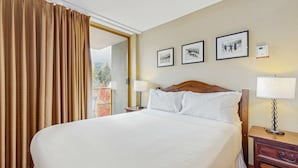Quarto de hotel com uma cama grande, duas mesas de cabeceira, um criado-mudo e uma janela com cortinas. Há três quadros na parede.