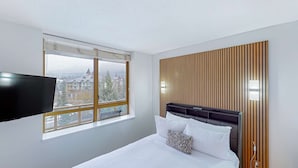 Une chambre d’hôtel avec un lit, un téléviseur fixé au mur et une fenêtre donnant sur des immeubles et des arbres.
