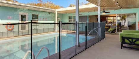 Un área de piscina exterior techada con techo de madera, cerramiento de vidrio y un sofá verde.