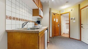 Eine kompakte Küche mit Spüle, Holzschränken und Mikrowelle. Eine Tür führt zu einem Badezimmer mit Dusche und Toilette.