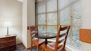 Ein Eckzimmer mit rundem Tisch, zwei Holzstühlen, einer Lampe und einem Fenster mit Jalousien.