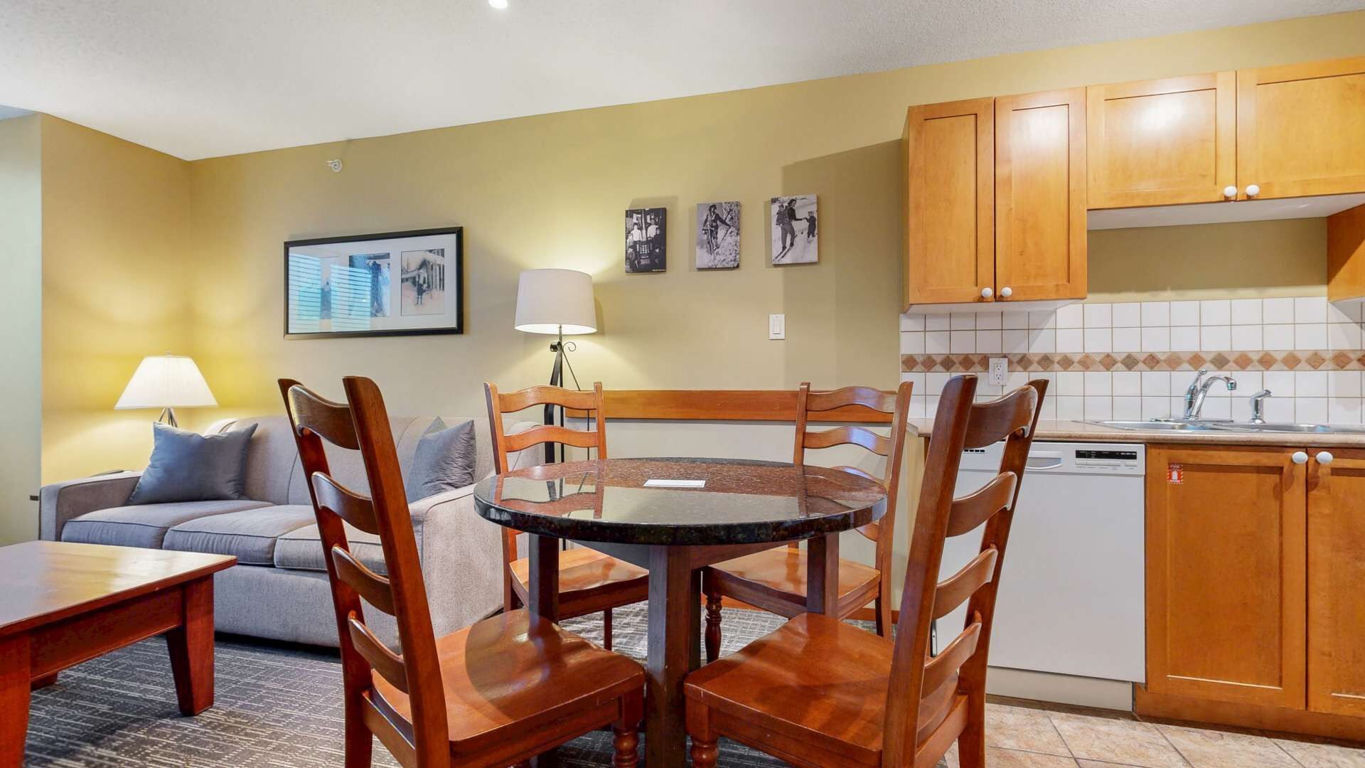 A compact living space with a round dining table, wooden chairs, a sofa, a lamp, and a kitchen area with wooden cabinets and a tiled backsplash.