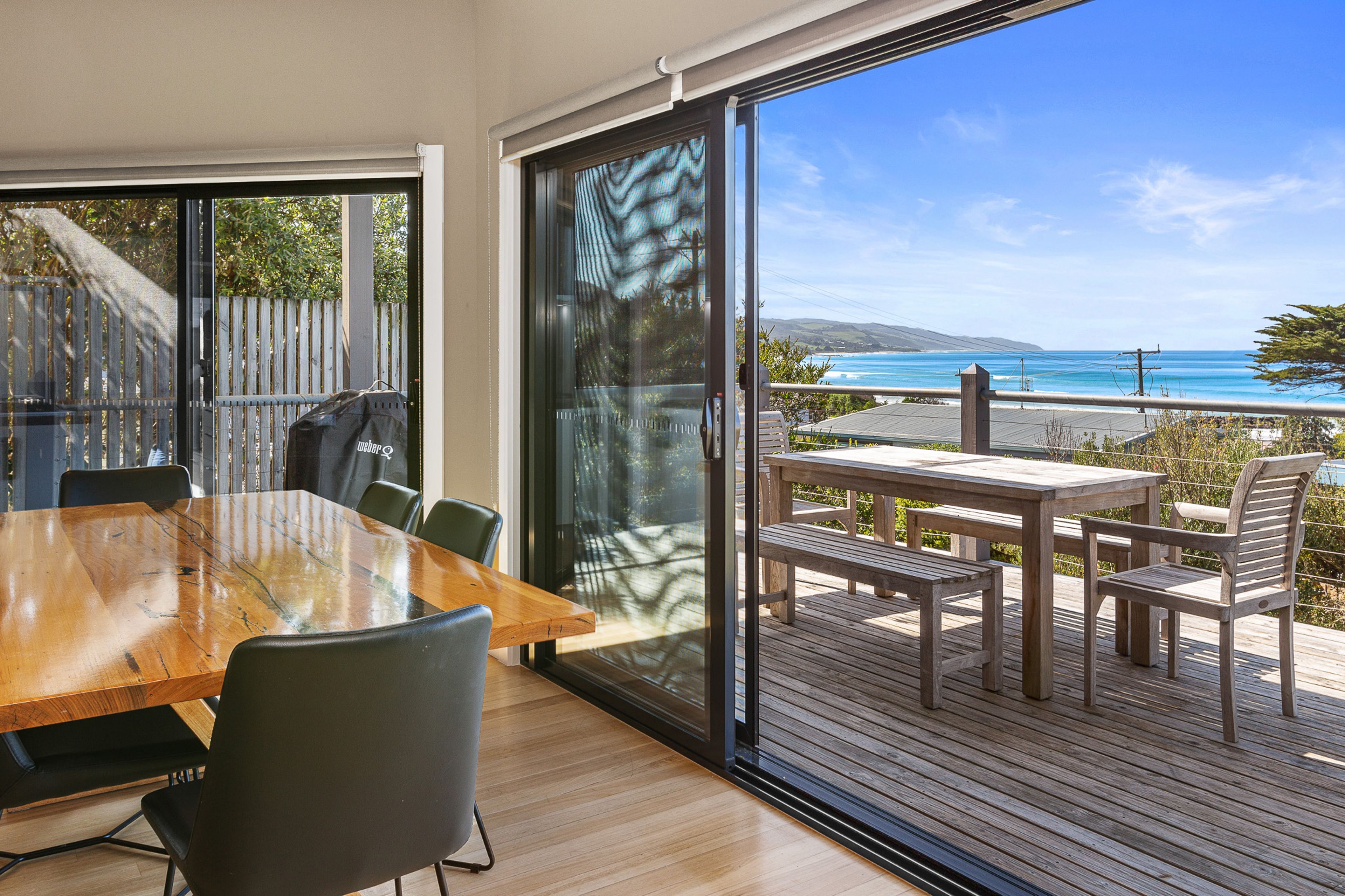A modern dining area with a wooden table and chairs, large sliding glass doors leading to a deck with a view of the ocean.