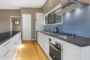 A modern kitchen with a stainless steel range, built-in oven, and a large window with a view of greenery. - Achilles (Apollo Bay)