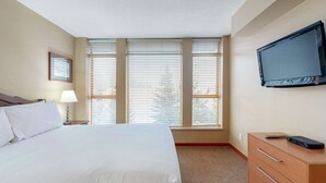 Une chambre d’hôtel avec un lit, un téléviseur fixé au mur, un bureau avec une lampe et une fenêtre avec des stores.