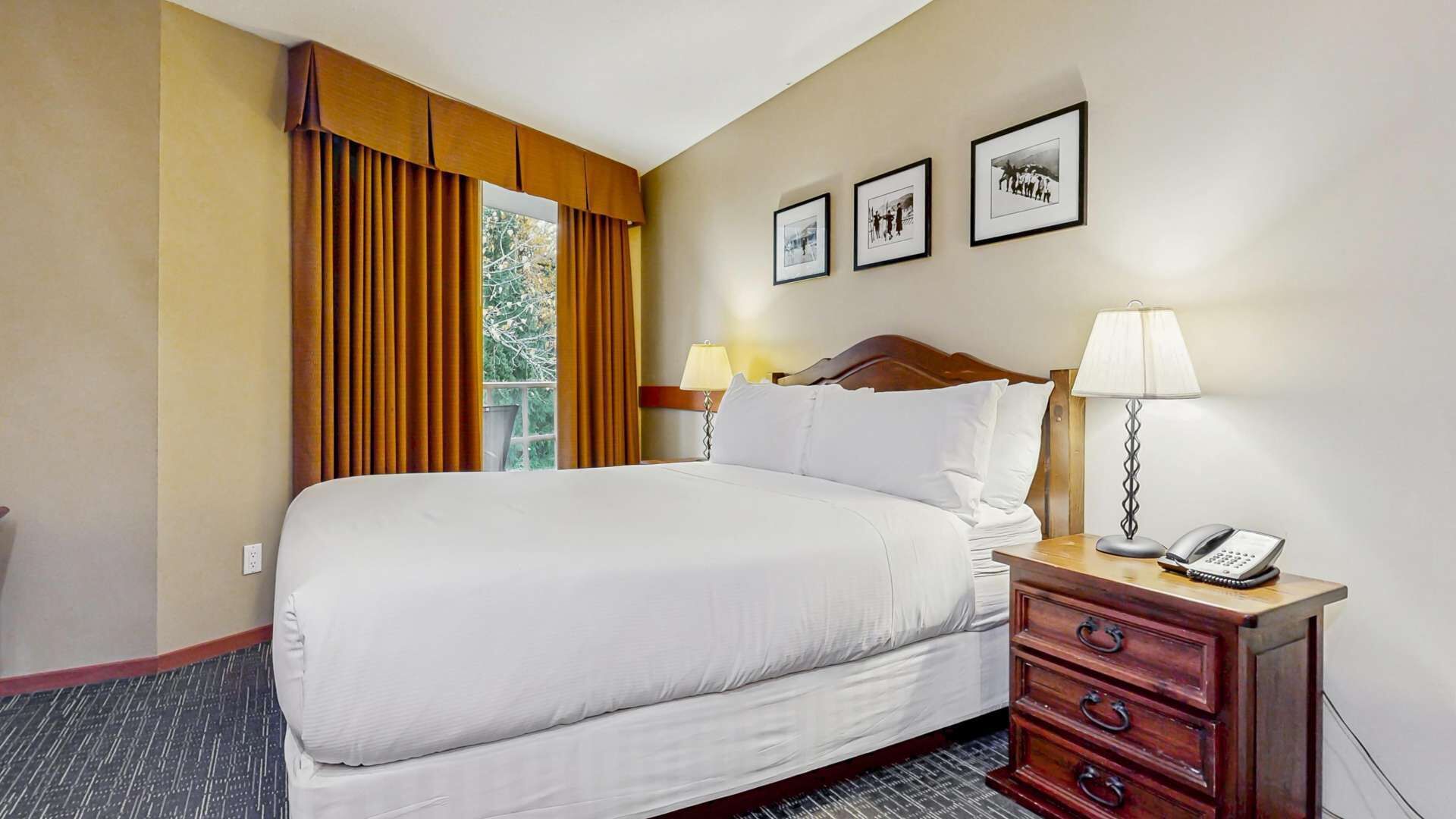 Un cuarto de hotel con una cama grande, una mesita de noche de madera con lámpara y teléfono, una ventana con cortinas, y tres cuadros en la pared.