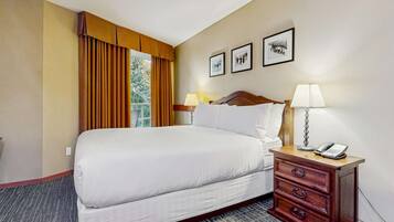 Un cuarto de hotel con una cama grande, una mesita de noche de madera con lámpara y teléfono, una ventana con cortinas, y tres cuadros en la pared.