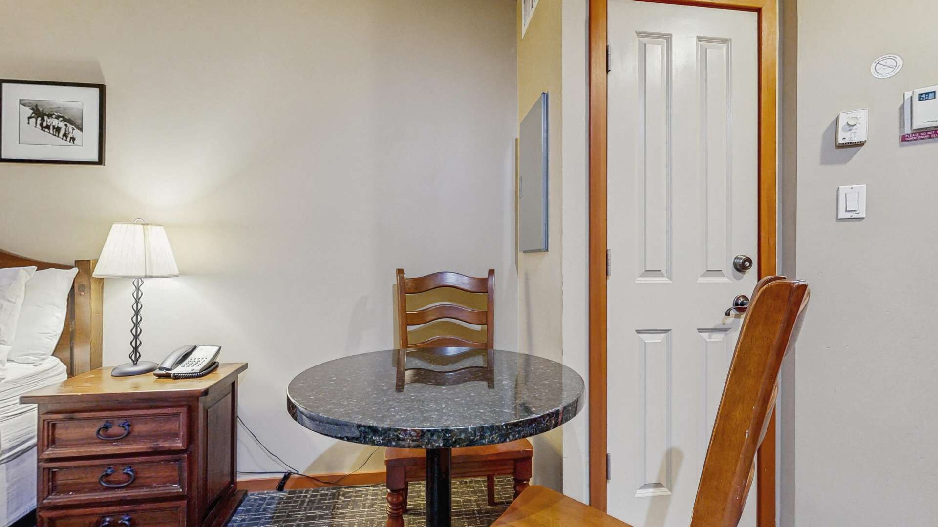 Una habitación de hotel con una mesa redonda de mármol, una silla de madera, un escritorio con lámpara y teléfono, y una cama con ropa blanca.