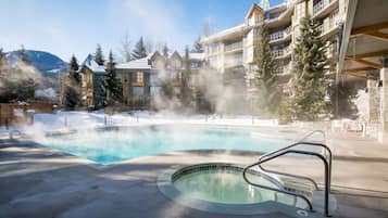 Un jacuzzi rodeado por una piscina, con un edificio que tiene balcones y un paisaje nevado al fondo.