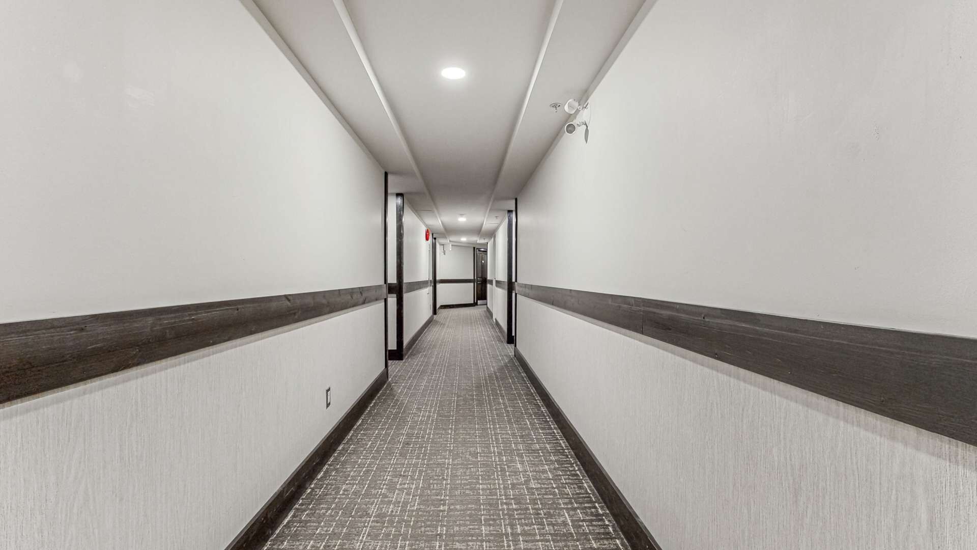 Un pasillo largo y estrecho con paredes blancas y un zócalo de madera oscura. El piso está cubierto de baldosas, y el pasillo está bien iluminado con luces del techo.