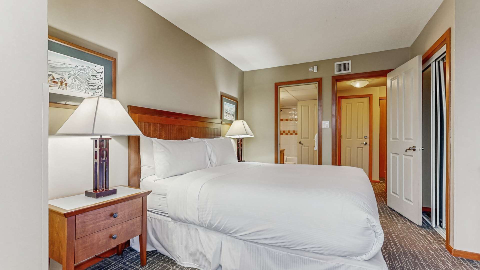 A hotel room with a large bed, two bedside tables with lamps, a wooden headboard, and a door leading to a bathroom.