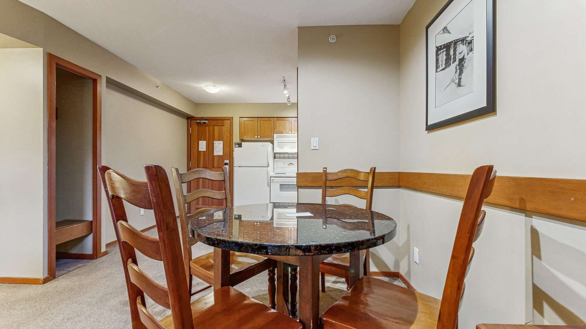 Un comedor con una mesa redonda y sillas de madera, una cocina al fondo y un cuadro en la pared.