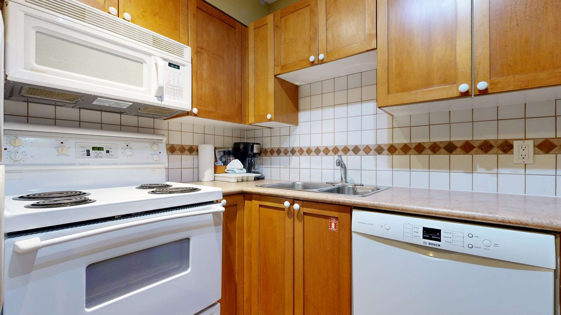 Een keuken met houten kastjes, een witte kookplaat, een magnetron, een vaatwasser en een betegelde spatwand.