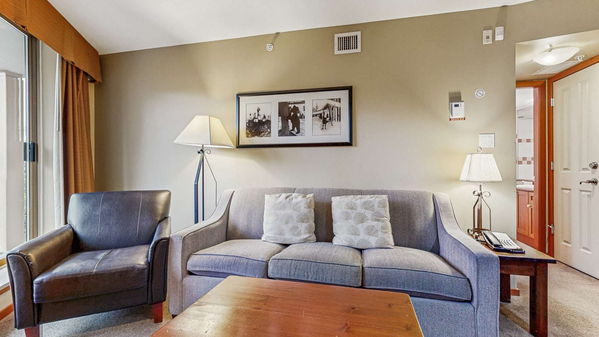 Una sala de estar con un sillón de cuero, un sofá gris y una mesa de centro de madera. Hay una foto enmarcada en la pared y una lámpara sobre una mesita auxiliar.