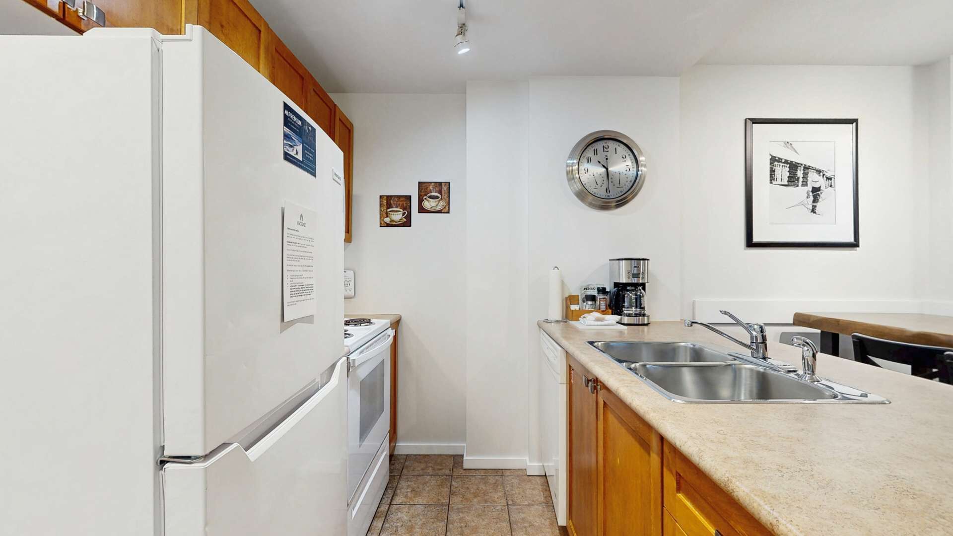 Una cocina moderna con refrigerador, estufa y lavaplatos. Hay armarios de madera, un reloj y un cuadro en la pared.