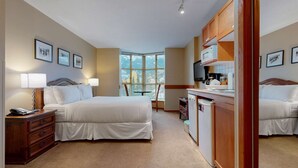 A hotel room with a large bed, a desk with a microwave, and a view of the city through large windows. - Whistler - Cascade Lodge 508 (Whistler)