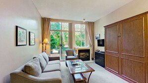 Ein Wohnzimmer mit Sofa, Sessel und Couchtisch. Ein Kamin, ein Fernseher und ein Kleiderschrank sind ebenfalls vorhanden.