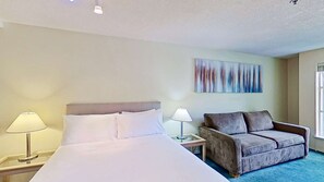 Une chambre d’hôtel avec un lit, deux tables de chevet avec des lampes, un canapé gris et un grand tableau abstrait accroché au mur.