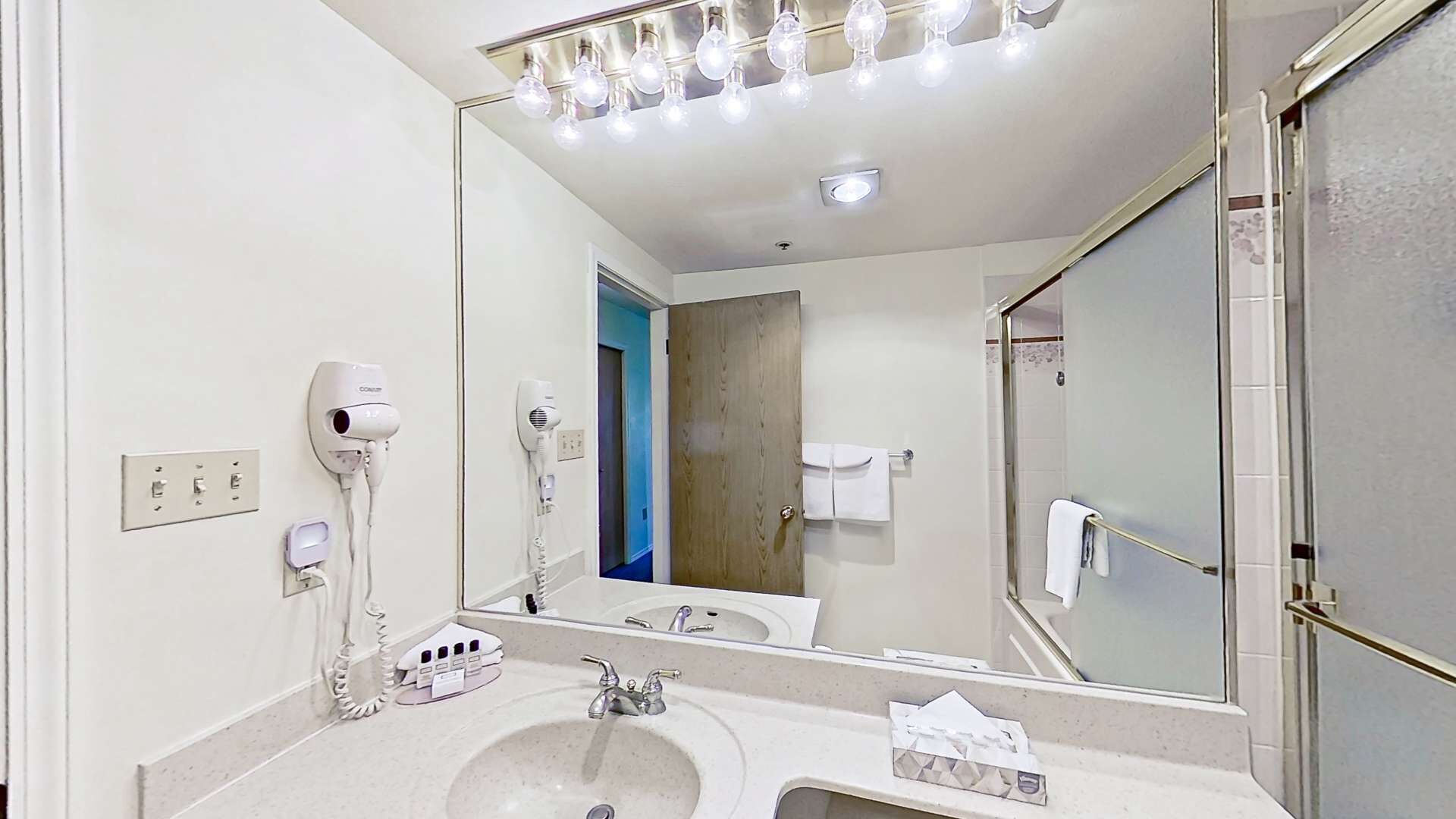 Une salle de bain d’hôtel avec un grand miroir, deux sèche-cheveux, un lavabo et un porte-serviettes.