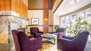 Un hall d’entrée avec une cheminée en pierre, des fauteuils recouverts d’un tissu violet, une table basse en verre et une grande fenêtre ornée de plantes.