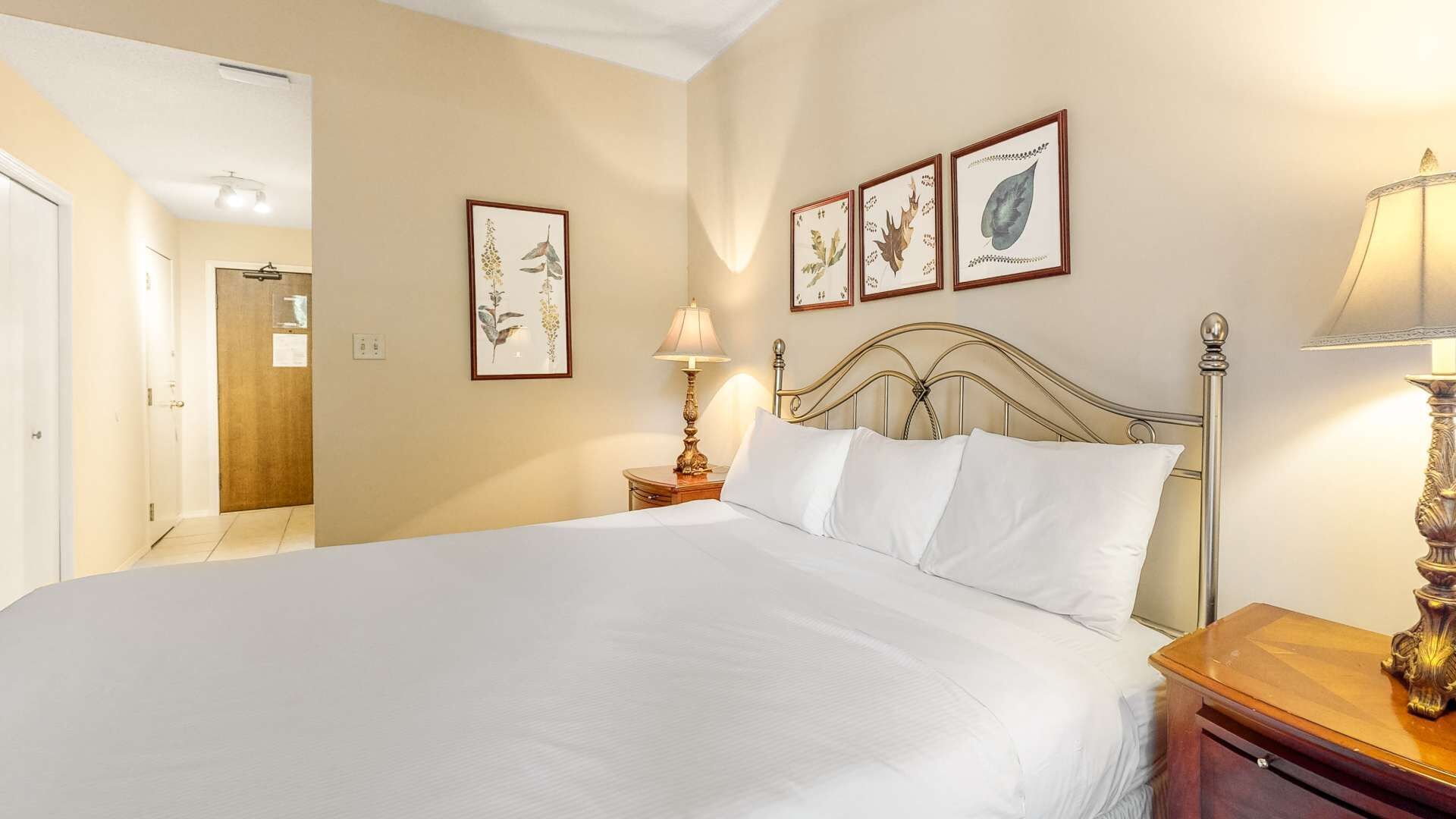A hotel room with a large bed, a wooden nightstand with a lamp, and three framed botanical prints on the wall.