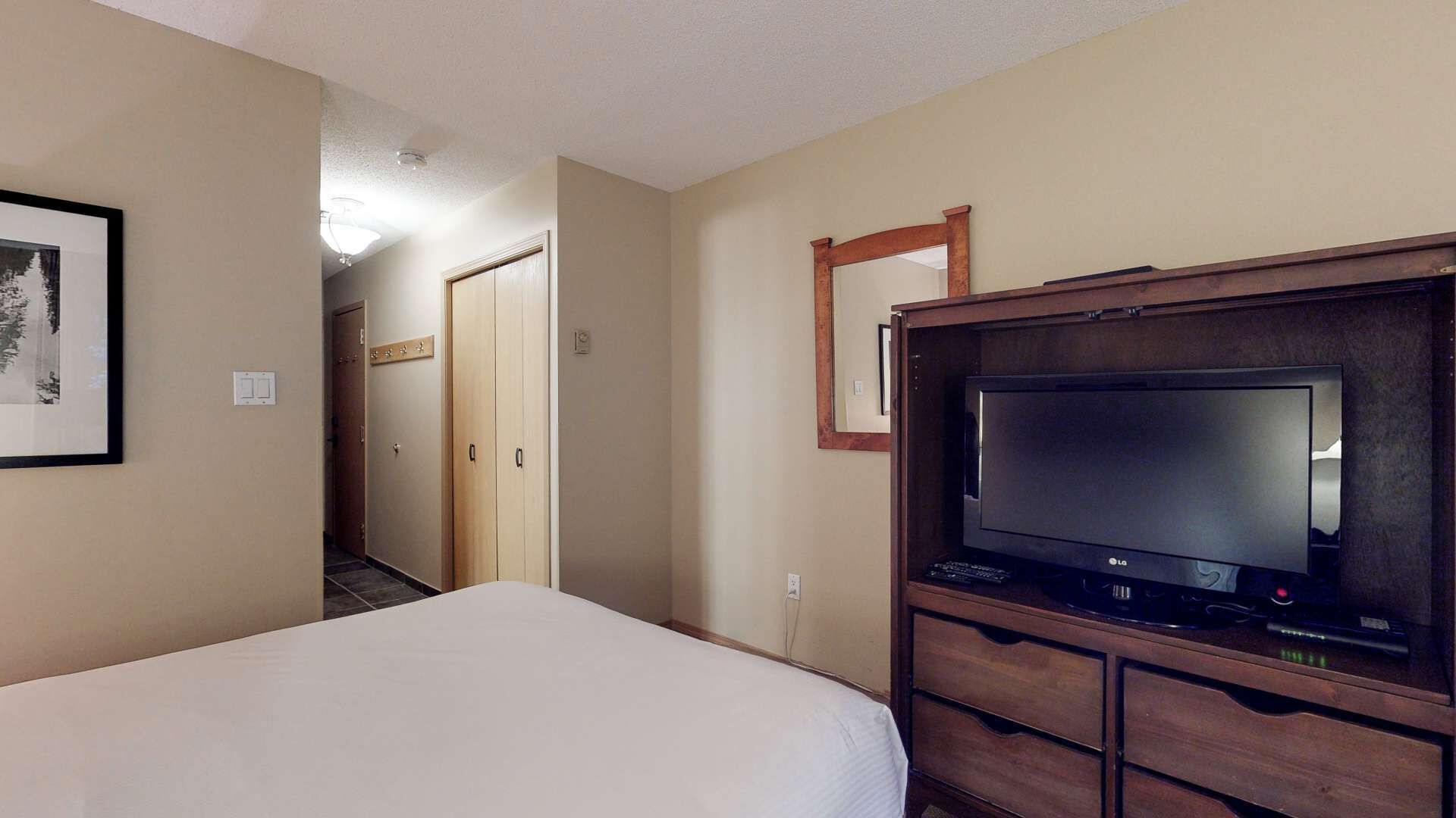 A hotel room with a bed, a TV on a wooden stand, a mirror, and a framed picture on the wall.