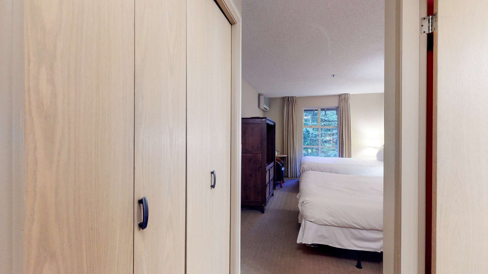 Una habitación de hotel con un armario de madera, una cama, un ventanal con cortinas y una mesita pequeña.