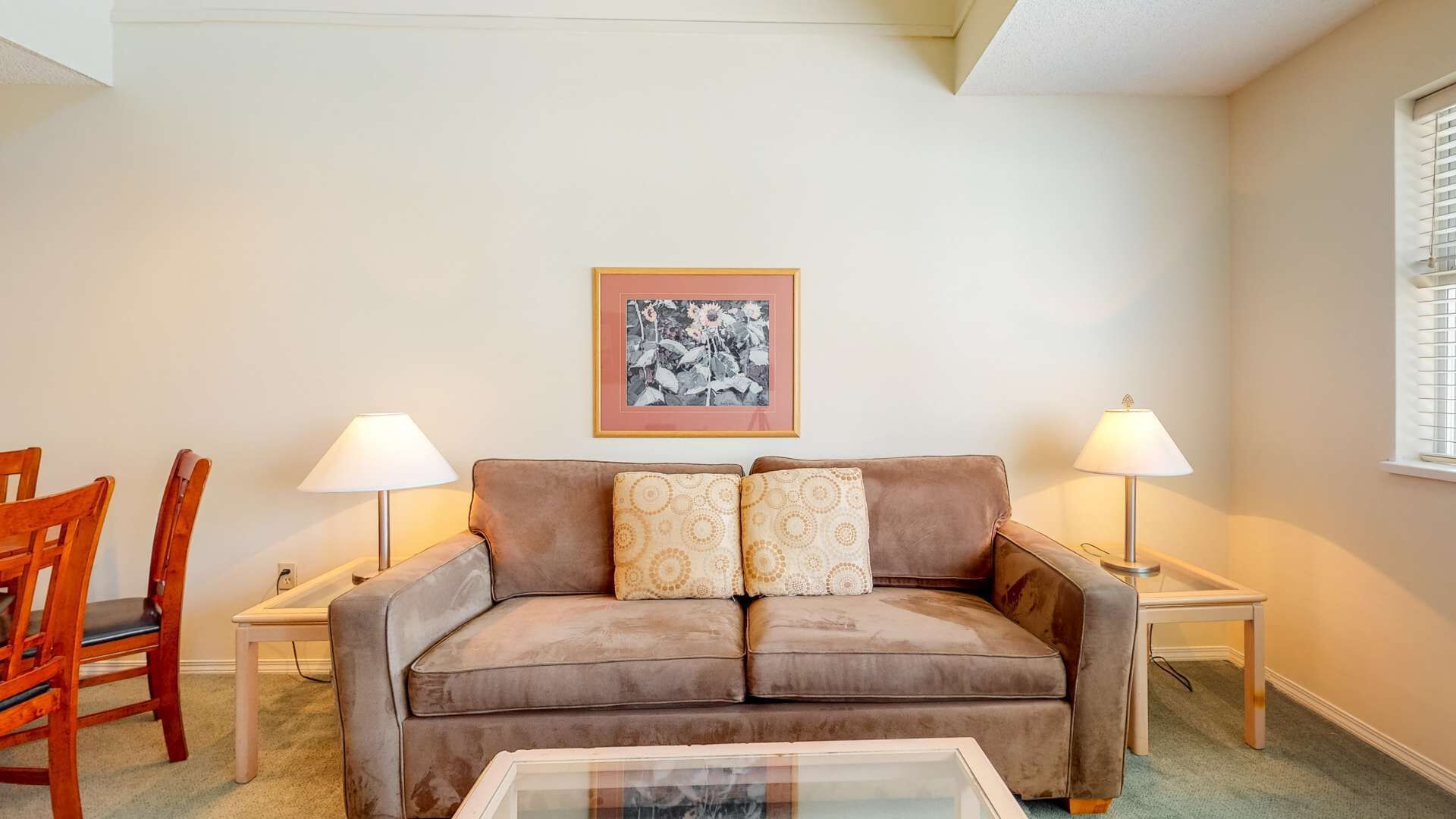 Ein Wohnzimmer mit braunem Sofa, zwei Lampen, einem gerahmten Bild an der Wand und einem Fenster mit Jalousien.