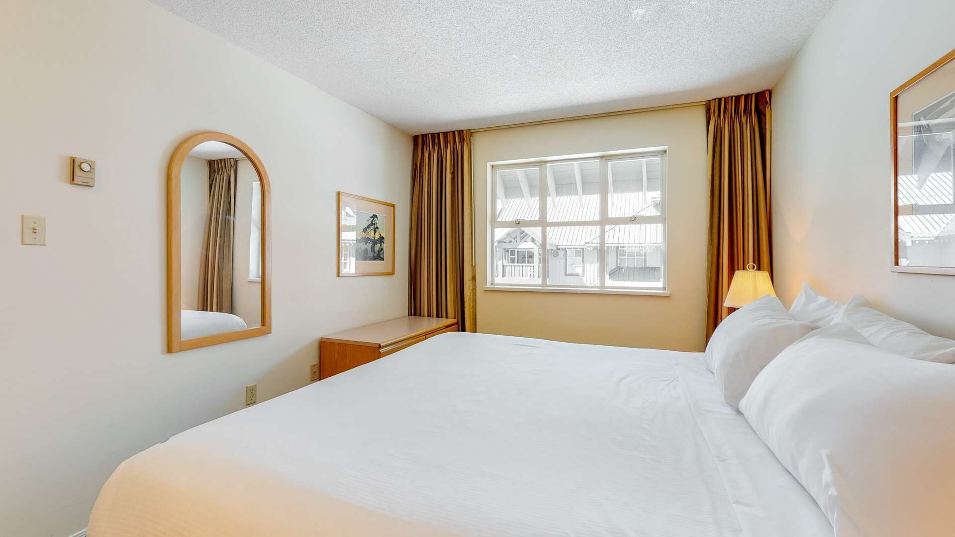 Une chambre d’hôtel avec un grand lit, une table de chevet, un miroir, une fenêtre avec des rideaux et un tableau encadré au mur.