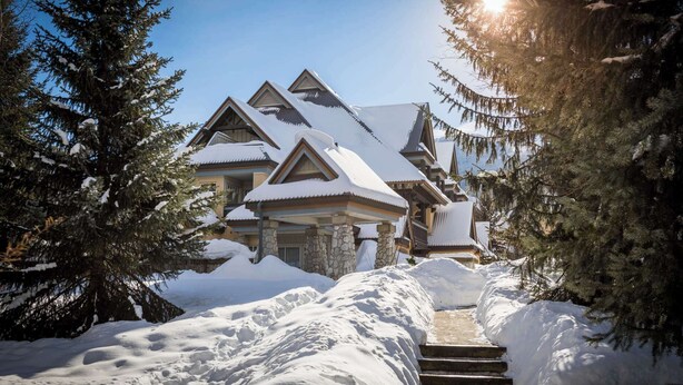 A snow-covered lodge with a stone entrance, multiple levels, and a prominent chimney.