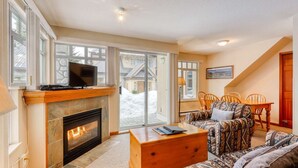 A cozy living room with a fireplace, a television on a mantel, a patterned sofa, and a dining area with chairs.