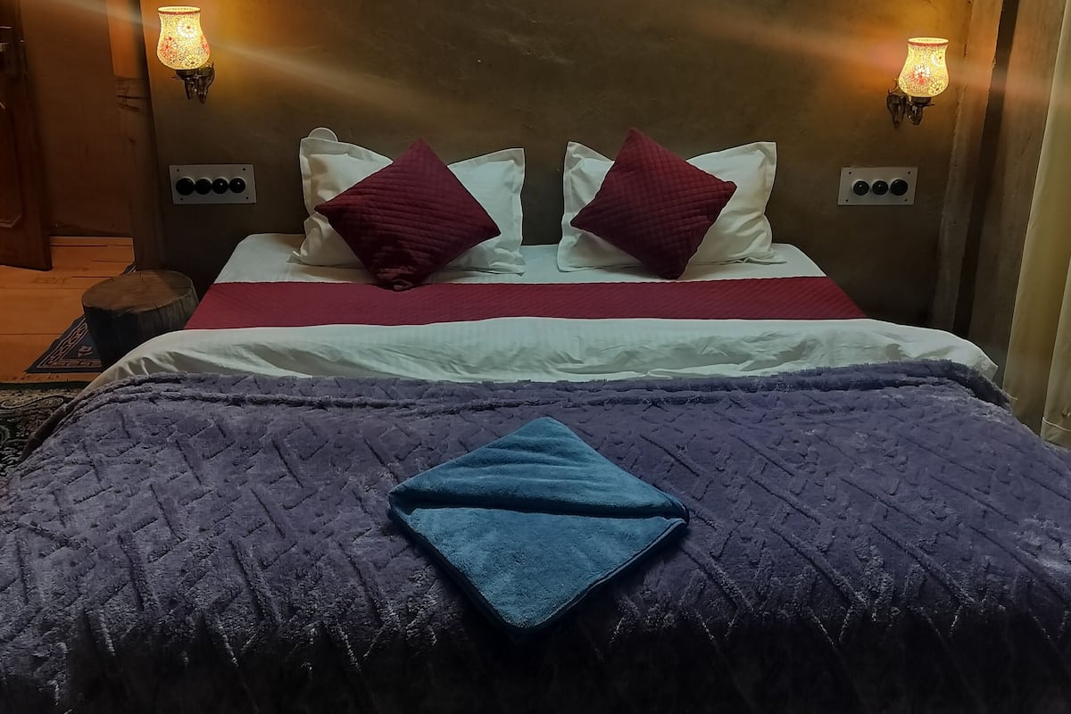 A bed with a purple blanket and two red pillows, a wooden headboard with a geometric pattern, and two wall-mounted lamps.