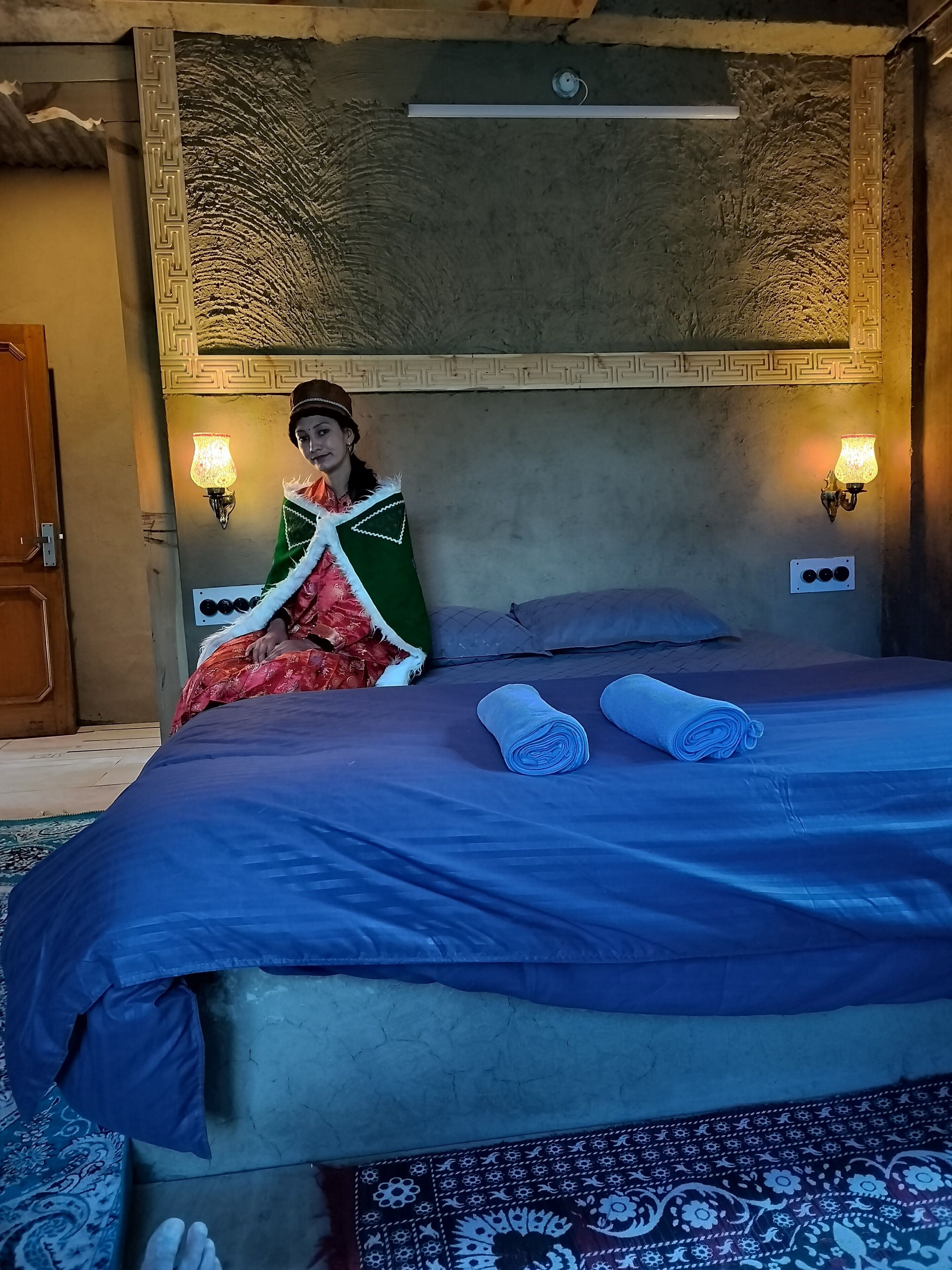 A bed with a blue bedspread and two rolled towels, a decorative wall with a geometric pattern, and two wall-mounted lamps.