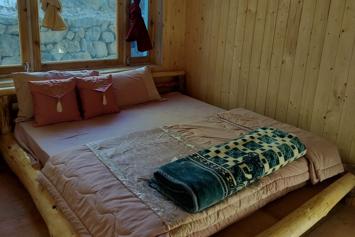 A wooden room with a bed, a window with curtains, and a stone wall outside.