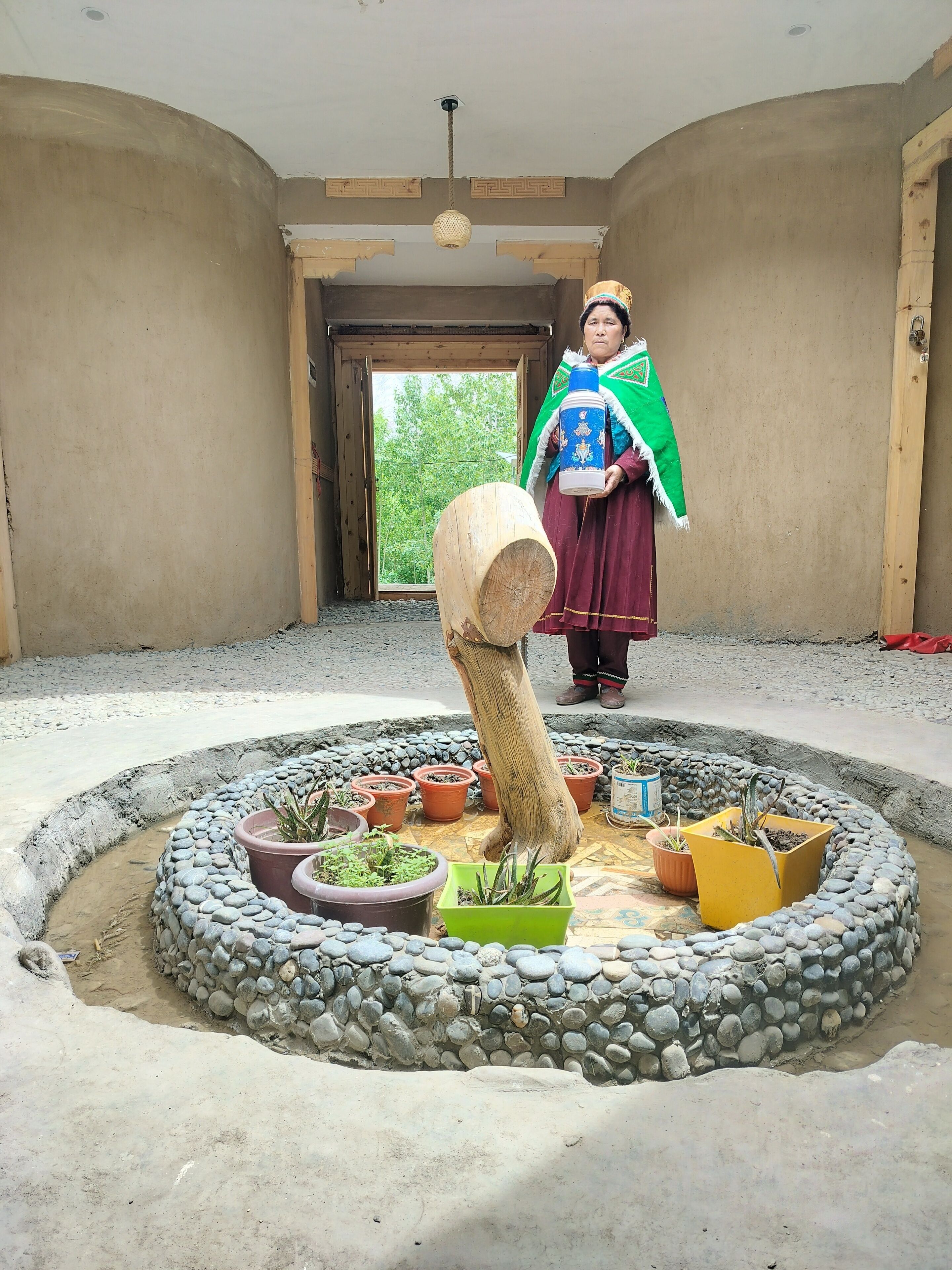 A room with a circular stone and plant arrangement, a wooden log, and a person holding a blue container.